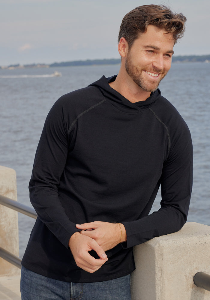 A man with short brown hair and a beard, wearing the Woolx Base Camp Hoodie in black, stands by a waterfront railing, smiling as he looks to the side. Calm water and a distant shoreline are visible in the background.